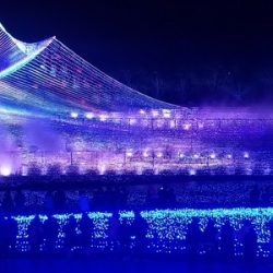 a mountain-like shape created of LED lights in all colors of the rainbow with fog surrounding it, along with two long blue tunnels of LED lights in the foreground, and several people walking around in front of them.