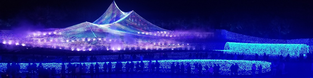a mountain-like shape created of LED lights in all colors of the rainbow with fog surrounding it, along with two long blue tunnels of LED lights in the foreground, and several people walking around in front of them.