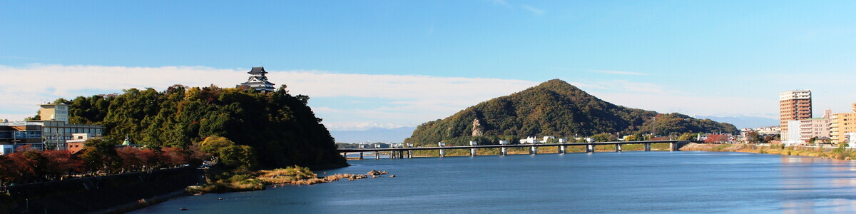 A wide river shot with Inuyama Castle shown at the top of a cliff on the left, and a mountain off in the distance in the middle with a bridge connecting the cliff to the land on the right side..