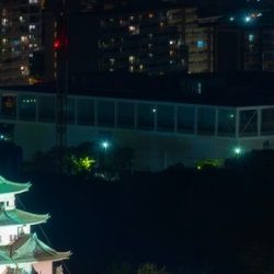 A night view of Nagoya Castle and its surrounding area from above. Green and white Nagoya Castle is underlit by bright white lights.