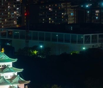 A night view of Nagoya Castle and its surrounding area from above. Green and white Nagoya Castle is underlit by bright white lights.