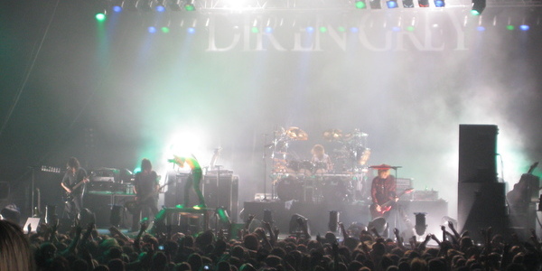 zoomed-out image of visual kei band Dir en grey performing on stage with blue and green lights shining down from above and the crowd below raising their arms