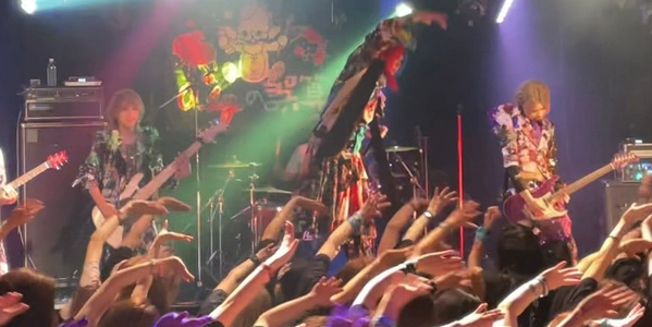image of visual kei band Royz's singer on stage moving his right arm over his head to the left side while all fans in front of him mimic him
