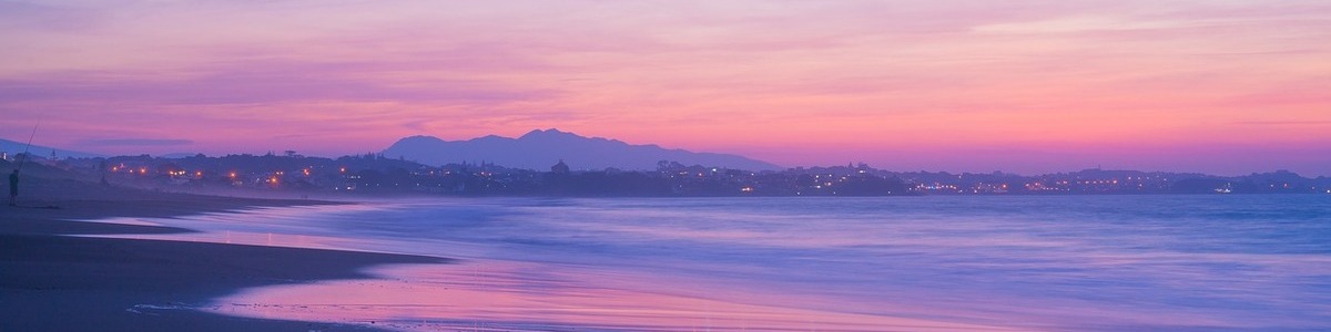 A purple-toned beach on Saku Island at sunset. The sky is filled with pinks and oranges, and the sand and water appear purple.